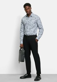 Langarmiges blaues Hemd mit schwarzem Paisleymuster, kombiniert mit schwarzen Hosen und schwarzen Schuhen, hält eine schwarze Lederaktentasche.