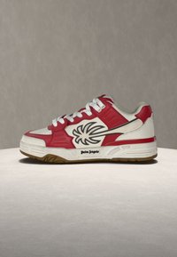 Λευκό και κόκκινο sneaker χαμηλού προφίλ με μαύρο λογότυπο φοίνικα και λευκά κορδόνια, τοποθετημένο πάνω σε ουδέτερη μπεζ επιφάνεια και φόντο.