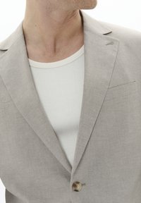 Világosszürke lenblézer egy gombbal, kerekített hajtókával és zsebbel. Fehér bordás póló fölött viselve.