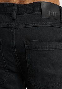 Jeans de mezclilla negros con un acabado texturizado, que presentan bolsillos traseros y un parche de cuero con el logotipo DEF en la cintura.