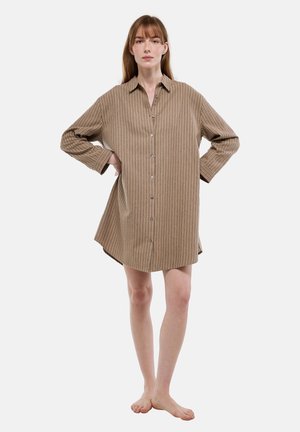 Femme debout pieds nus portant une robe chemise ample à manches longues, rayée marron avec des boutons, les mains sur les hanches, sur un fond blanc uni.