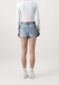 Osoba stojící zády, oblečená v bílém dlouhém rukávu crop topu, potrhlých světle modrých denimových šortkách, bílých ponožkách a bílých teniskách.