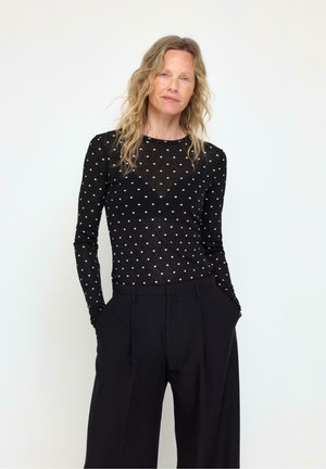 Kvinde med langt blondt hår iført en sort gennemsigtig polkaprikket bluse med lange ærmer og sorte højttaljede bukser med vide ben og hænderne i lommerne.