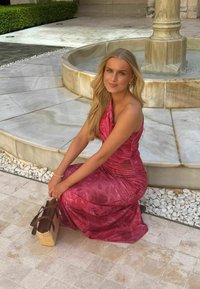 Νεαρή γυναίκα με μακριά ξανθά μαλλιά σε κόκκινο φόρεμα με λουλούδια, καθισμένη σε πέτρινο πάτωμα δίπλα σε μαρμάρινο σιντριβάνι με μια υφαντή τσάντα δίπλα της.