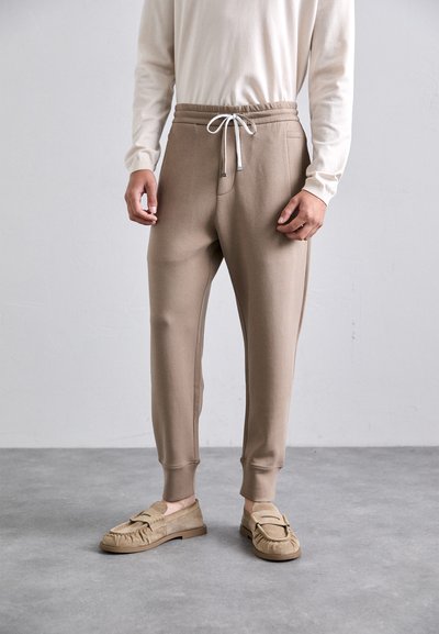 Uomo che indossa pantaloni jogger marrone chiaro con cordino bianco e mocassini beige, in piedi su un pavimento grigio contro un muro semplice.