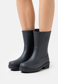 Bottes de pluie noires en caoutchouc mi-mollet portées sur des jambes nues avec des semelles texturées, présentées sur un fond blanc uni.