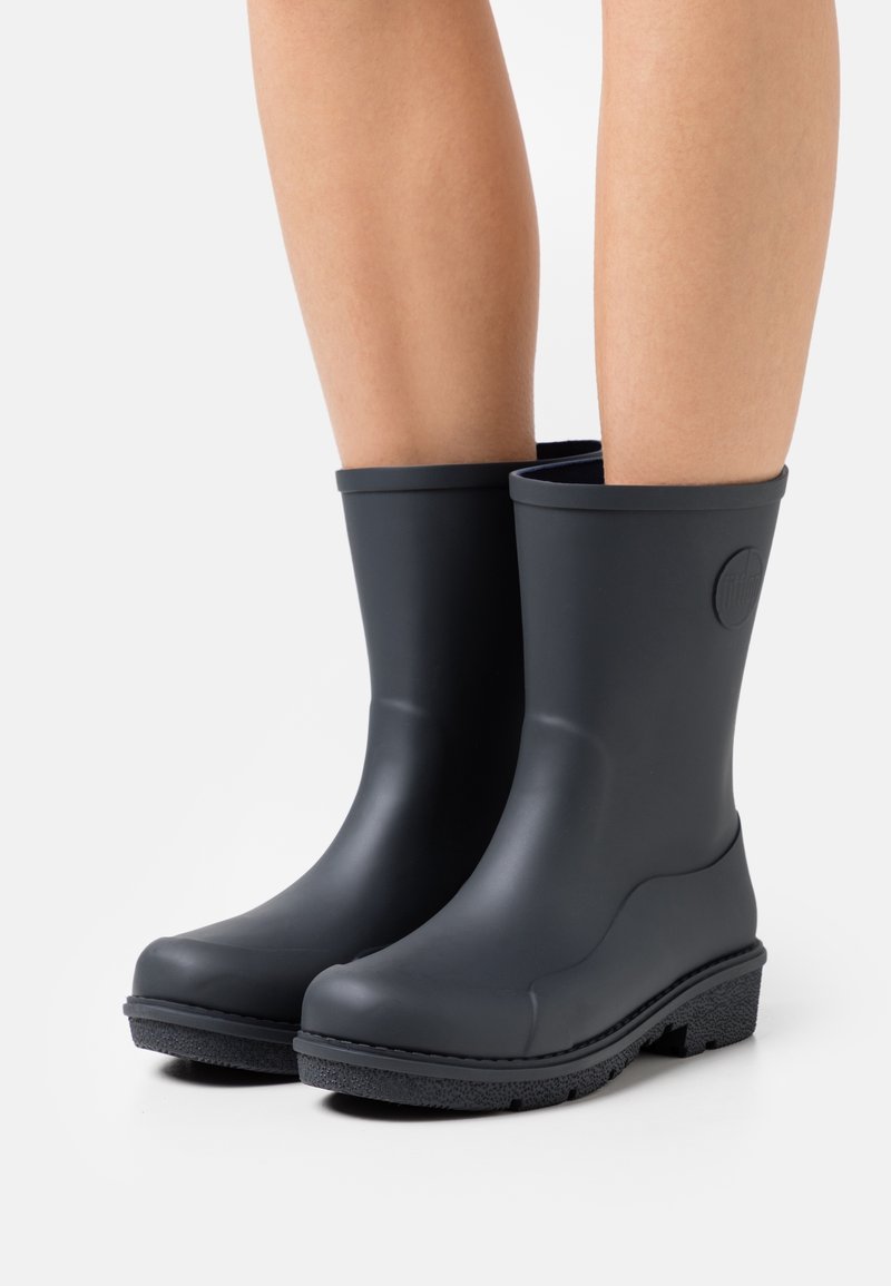 Bottes de pluie noires en caoutchouc mi-mollet portées sur des jambes nues avec des semelles texturées, présentées sur un fond blanc uni.