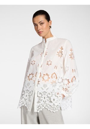 Frau trägt eine weiße langärmelige Bluse mit floraler Cut-Out-Stickerei und Stehkragen, kombiniert mit hellgrauen Hosen.