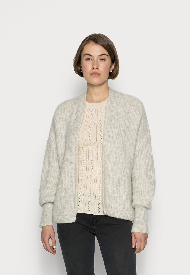 Šviesiai pilkas vilnonis megztinis su ilgomis rankovėmis, atviromis priekinėmis dalimis ir minkšta tekstūra, uždėtas ant banguotos grietinėlės spalvos megztinio ir tamsių kelnių.