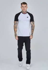 White T-shirt with black raglan sleeves, featuring a small logo on the chest, paired with black jeans and white sneakers.
