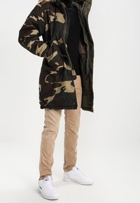 Camouflage insulated parka jacket with a hood, paired with beige slim-fit pants and white sneakers, featuring a smooth, shiny fabric.