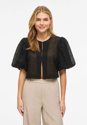 Młoda kobieta w czarnej koronkowej krótkiej bluzce z bufkami na rękawach i beżowych spodniach z wysokim stanem, stojąca na tle białego tła.