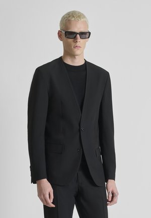 Giovane uomo con capelli biondi corti, che indossa occhiali da sole neri e una giacca da abito nera sopra una camicia nera, su uno sfondo grigio uniforme.