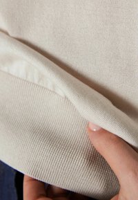 Main tenant et soulevant le bord d'un tissu tricoté beige, montrant la texture et le détail de l'ourlet sur un fond de vêtements foncés.