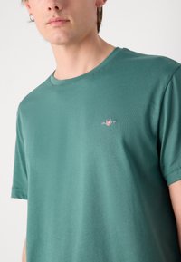 Puuvillane t-särk türkiissinisena, koos ringikujulise kaelusega. Vasakul rinnal on väike tikandiga logo. Sile tekstuur ja lühikesed varrukad.