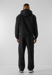 Mężczyzna stoi tyłem, ubrany w czarną bluzę z kapturem, czarne spodnie dresowe i białe trampki na jednolitym szarym tle.