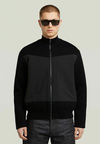 Giacca nera con zip, parte superiore in maglia testurizzata e parte inferiore in tessuto liscio. Caratterizzata da un colletto alto e due tasche sul petto. Indossata con occhiali da sole scuri.