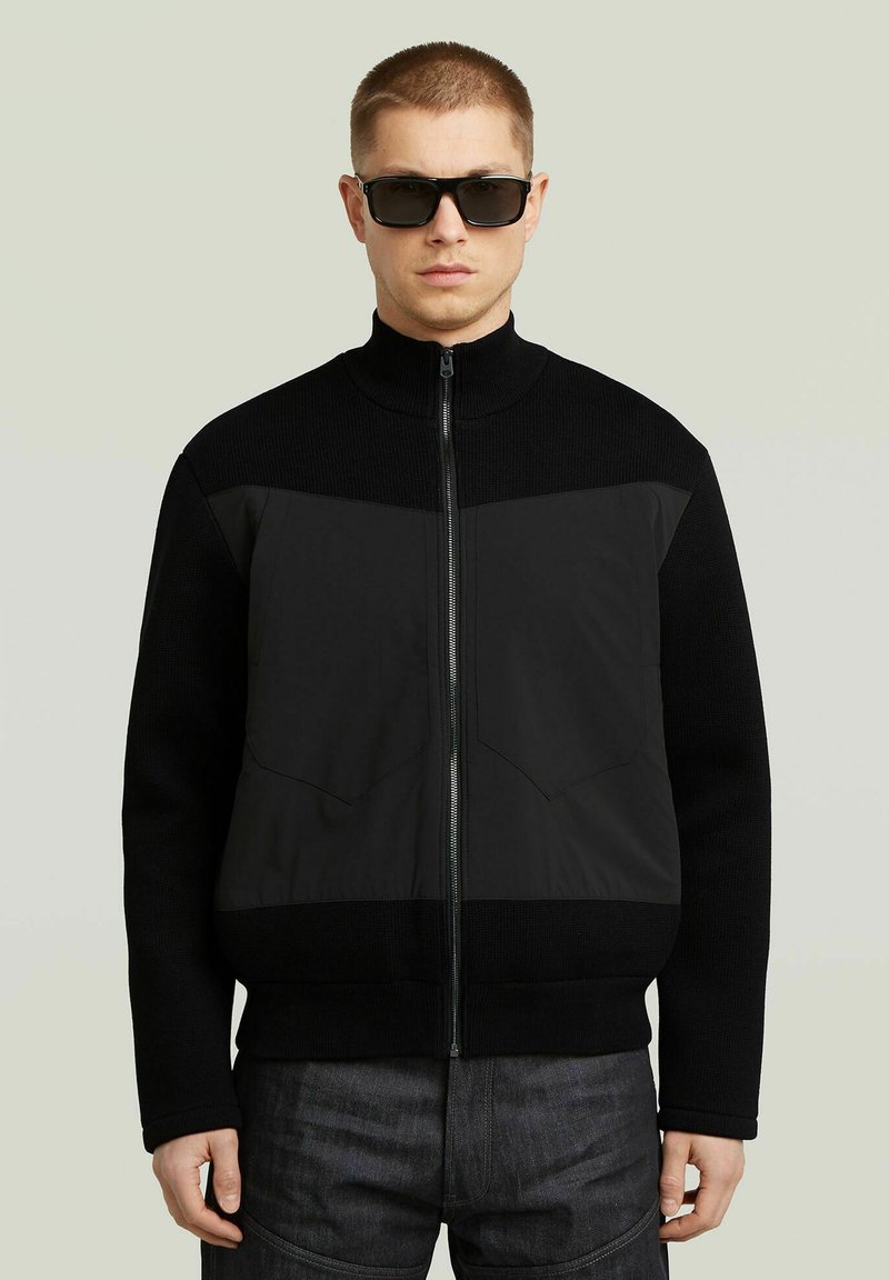 Giacca nera con zip, parte superiore in maglia testurizzata e parte inferiore in tessuto liscio. Caratterizzata da un colletto alto e due tasche sul petto. Indossata con occhiali da sole scuri.