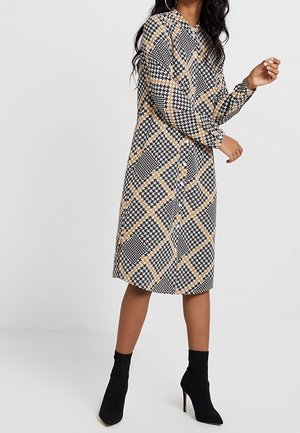 Frau trägt ein knielanges Kleid mit langen Ärmeln, schwarzen, weißen und beigen geometrischen Mustern und schwarze hochhackige Stiefeletten.
