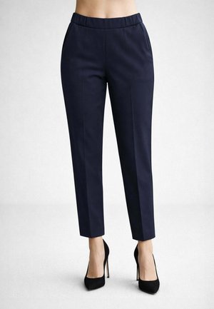 Donna che indossa pantaloni sartoriali blu navy alla caviglia e tacchi alti neri, in piedi davanti a uno sfondo grigio chiaro uniforme.