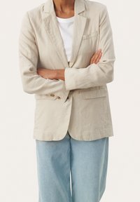 Vaaleanbeige pellavapuku, jossa on viistetyt läpät, kahden napin suljenta ja sivutaskut, kerrostettuna valkoisen t-paidan kanssa, joka on yhdistetty rentoihin farkkuihin.