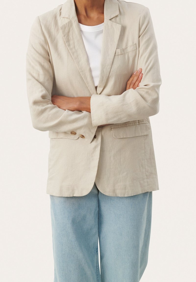 Vaaleanbeige pellavapuku, jossa on viistetyt läpät, kahden napin suljenta ja sivutaskut, kerrostettuna valkoisen t-paidan kanssa, joka on yhdistetty rentoihin farkkuihin.