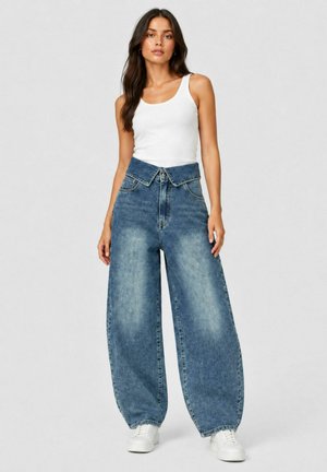 Vrouw draagt een wit hemdje, hooggetailleerde wijdere blauwe jeans met opgevouwen tailleband en witte sneakers, staand tegen een effen achtergrond.