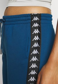 Blå joggingsbyxor med elastisk midjeband och snörning. Har svarta sidpaneler med vitt logomönster. Mjuk textur och avslappnad passform.