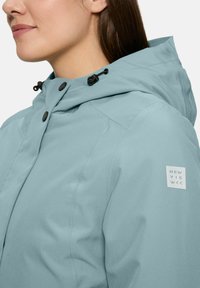 Vrouw in een lichtblauwe hooded jack met zwarte knopen en een "New View" patch op de linkermouw.