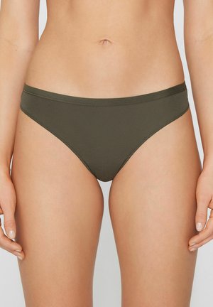 Frau trägt schlichte olivgrüne Bikini-Unterwäsche, gezeigt von der Taille bis zur Mitte des Oberschenkels vor hellem Hintergrund.