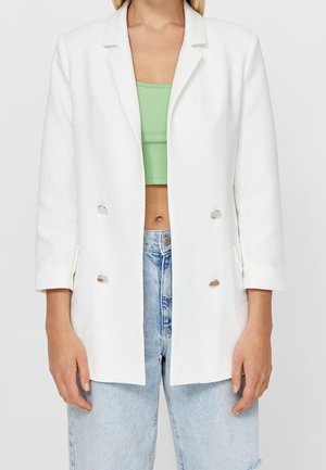 Femme portant un blazer blanc croisé sur un débardeur court vert et un jean taille haute bleu clair.