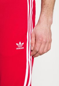 Červené sportovní kalhoty s b ílými bočními pruhy a vyšitým bílým logem Adidas. Materiál vypadá hladce a má přiléhavý střih.