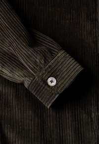 Olive zielony mankiet koszuli z korduroju z pionową fakturą prążków oraz białym guzikiem z czterema otworami.