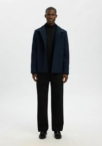 Marineblå uldblandingsfrakke med spidsrevers, knaplukning og tekstureret finish; parret med en sort turtle-neck og løse sorte bukser.