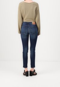 Persona vistiendo una blusa de manga larga beige, jeans ajustados azul oscuro con bolsillos traseros y sandalias de tacón negras, de pie contra un fondo blanco liso.