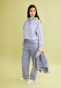 Lichtblauwe cropped hoodie met een trekkoord, gecombineerd met losse lichtblauwe jeans. Witte instappers en een bijpassende denimjas. Groene achtergrond.