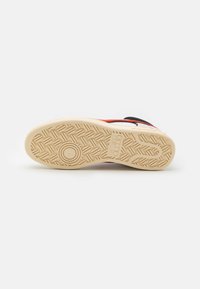 Suola di sneaker in gomma beige chiaro, dotata di un motivo testurizzato con design circolari e lineari. Accenti in rosso e nero lungo il bordo superiore.