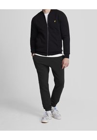 Sort zip-trøje med ribbede manchetter, parret med sorte tætsiddende bukser. Hvid t-shirt under. Grå sneakers med blå accenter.