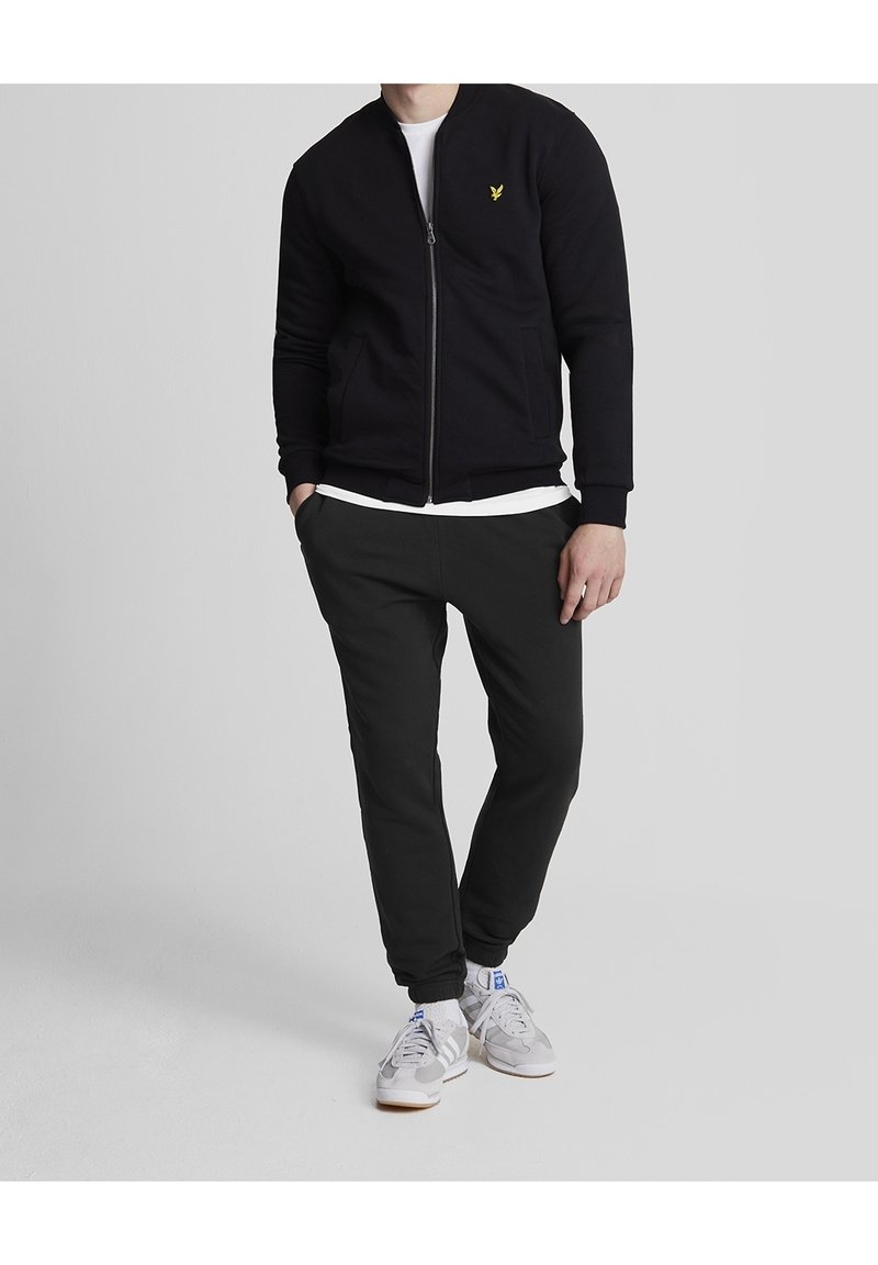 Sort zip-trøje med ribbede manchetter, parret med sorte tætsiddende bukser. Hvid t-shirt under. Grå sneakers med blå accenter.