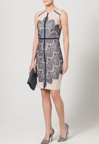 Beige bezrukávové šaty s tmavomodrou krajkovou vrstvou, s centrálním zipem a vertikálními švy. V kombinaci s černou kabelkou a stříbrnými podpatky.