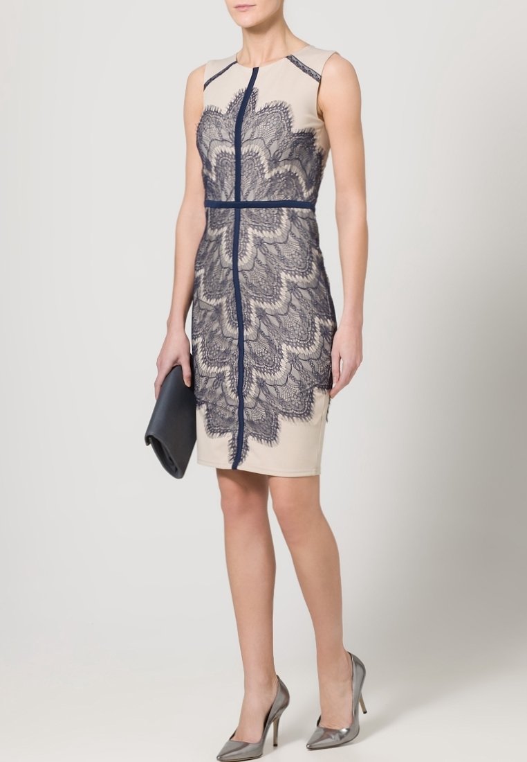 Beige bezrukávové šaty s tmavomodrou krajkovou vrstvou, s centrálním zipem a vertikálními švy. V kombinaci s černou kabelkou a stříbrnými podpatky.