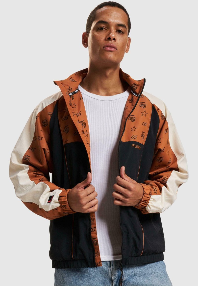 Farveblokkeret jakke med brune, sorte og cremefarvede paneler. Designet inkluderer gentaget branding, elastiske manchetter og en ståkrave.