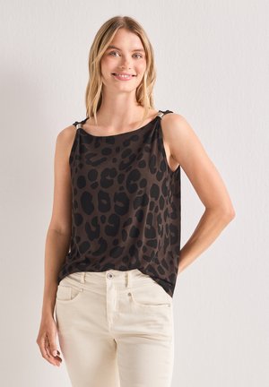 Jeune femme aux cheveux blonds portant un haut sans manches imprimé léopard marron foncé et noir et un pantalon beige clair, souriante avec une main dans le dos.