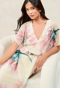Vestido maxi floral con escote en V, mangas cortas y una silueta fluida. Presenta colores pastel, acentos de lentejuelas y un patrón inspirado en jardines.