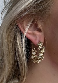 Gouden creolen met ingewikkelde clusters van heldere edelstenen, een gestructureerd oppervlak en een gedraaide vorm, gedragen in het oor.