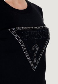 Maglione nero lavorato a maglia con un patch ricamato a forma triangolare decorato con strass, che mostra il logo "GUESS" e dettagli testuali.