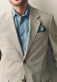 Blazer de cuadros claros hecho de un tejido texturizado, con dos botones y un pañuelo de bolsillo. Debajo, se puede ver una camisa de chambray azul.