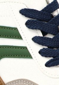 Zapatilla blanca con cordones azul oscuro y dos franjas verdes paralelas cosidas en el lateral.