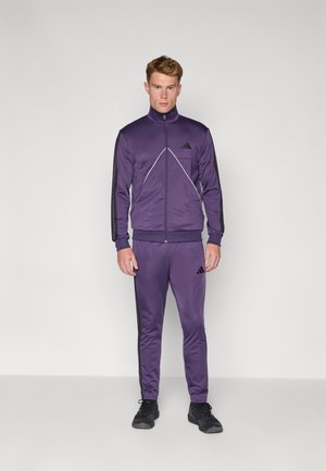 Entrenador morado hecho de tejido suave, con chaqueta de cremallera con franjas negras y pantalones a juego. Logo de Adidas visible en ambos.