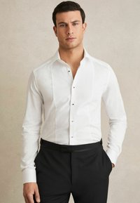 Cămașă albă cu un panou frontal texturat, nasturi negri și un guler clasic, purtată cu pantaloni negri. Țesătură fină, croială ajustată.